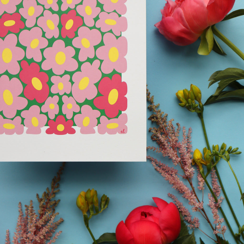 Close up of a printed artwork featuring a whimsical pattern of wobbly pink daisies on a blue background, displayed against a backdrop of pink flowers and green foliage.