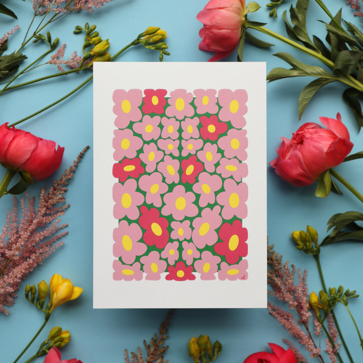 A printed artwork featuring a whimsical pattern of wobbly pink daisies on a blue background, displayed against a backdrop of pink flowers and green foliage.