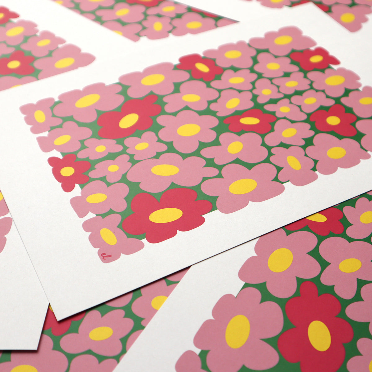 A printed artwork featuring a whimsical pattern of wobbly pink daisies on a blue background, displayed overlapping 
